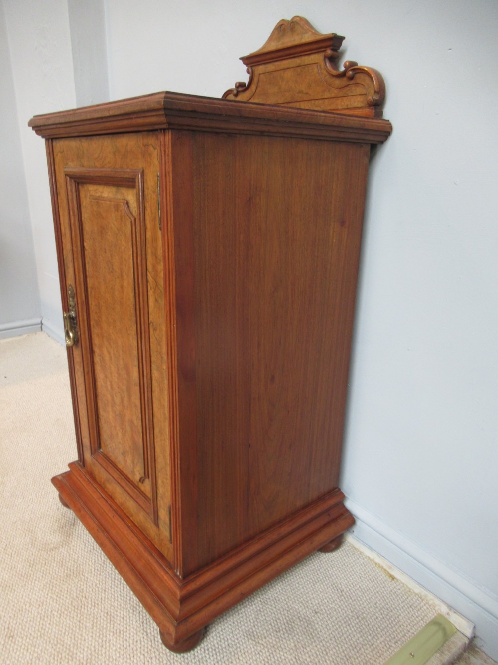 A late Victorian burr walnut bedside cabinet featuring a single panelled door with a brass drop handle, a shaped pediment gallery, and a plinth base on bun feet. View V