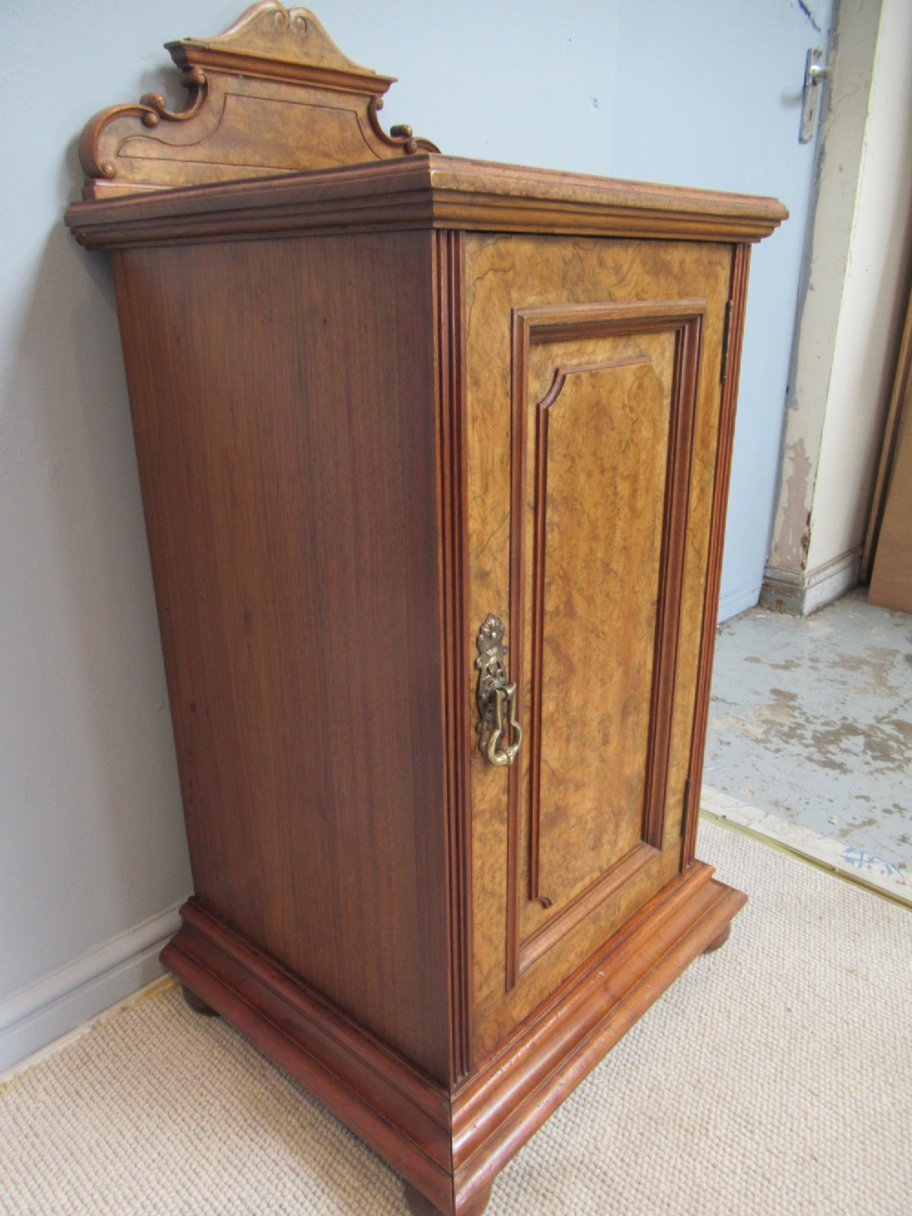 A late Victorian burr walnut bedside cabinet featuring a single panelled door with a brass drop handle, a shaped pediment gallery, and a plinth base on bun feet. View VI
