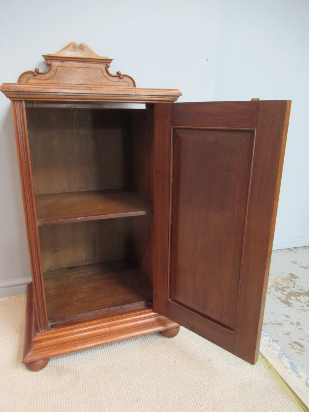 A late Victorian burr walnut bedside cabinet featuring a single panelled door with a brass drop handle, a shaped pediment gallery, and a plinth base on bun feet. View VII
