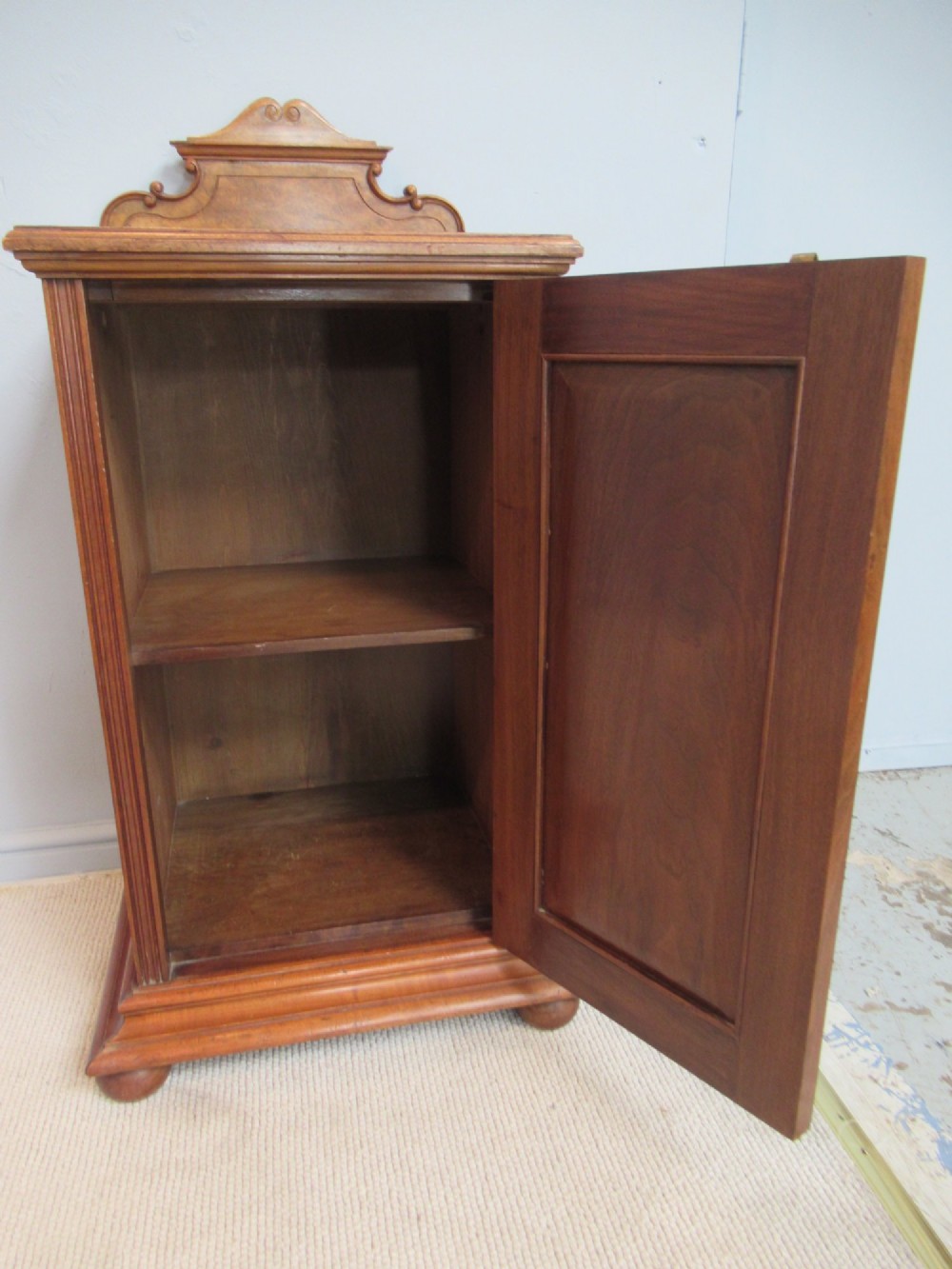 A late Victorian burr walnut bedside cabinet featuring a single panelled door with a brass drop handle, a shaped pediment gallery, and a plinth base on bun feet. View VIII