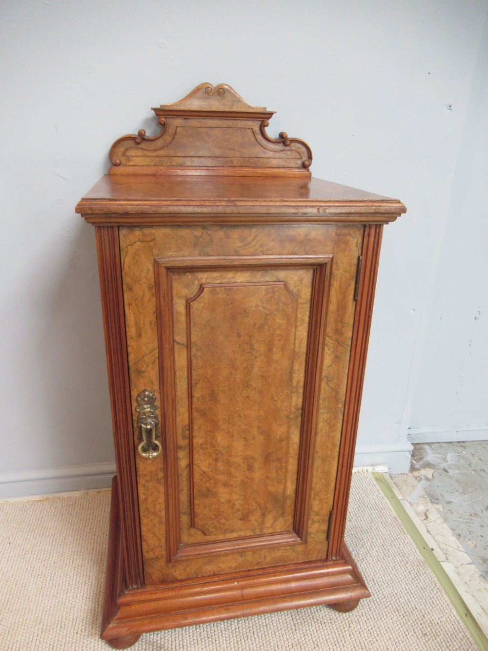 A late Victorian burr walnut bedside cabinet featuring a single panelled door with a brass drop handle, a shaped pediment gallery, and a plinth base on bun feet. View IX