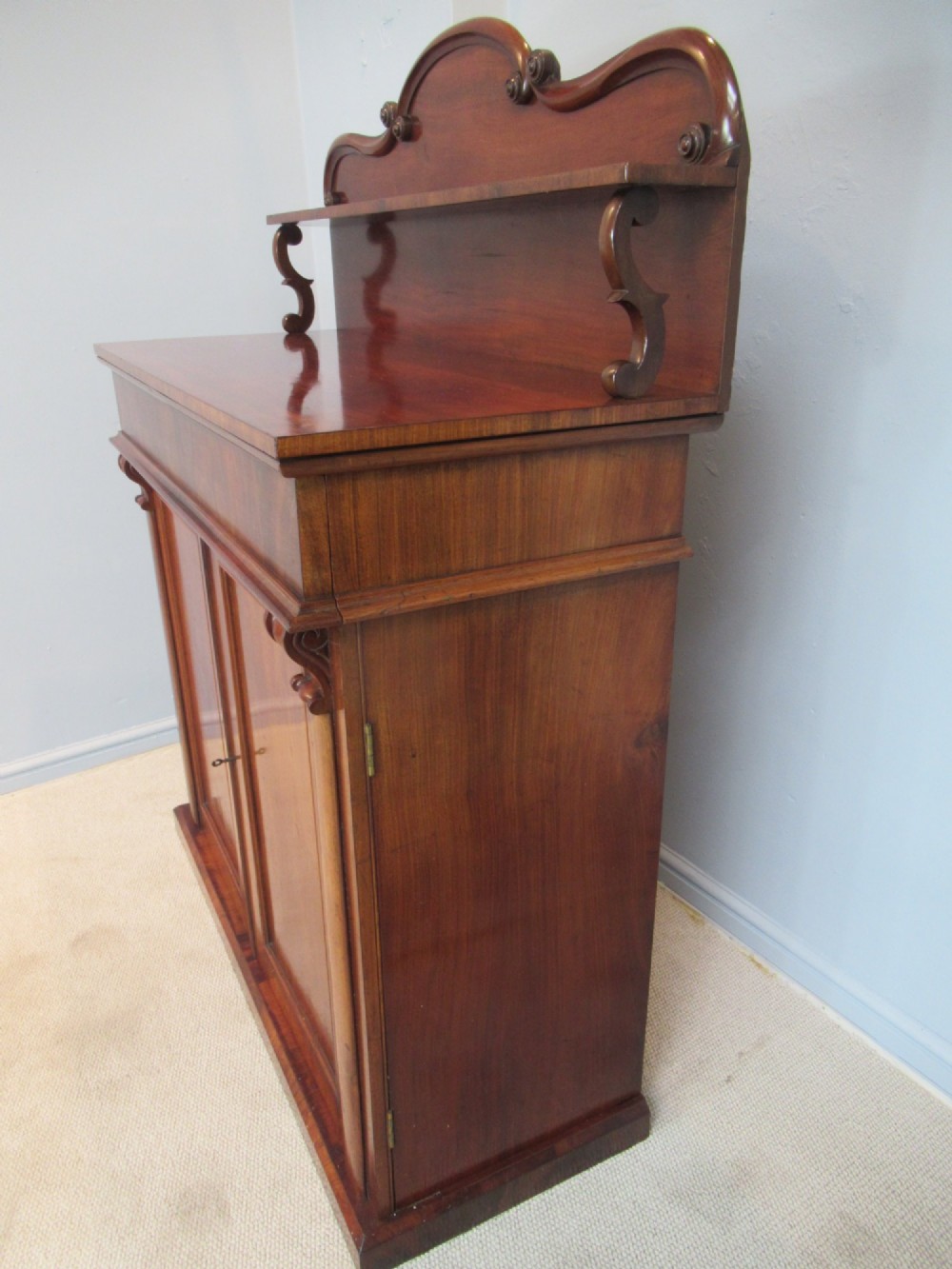 A Victorian mahogany chiffonier with a raised gallery shelf supported by carved scrolls above a two-door cabinet on a solid plinth base. View IX