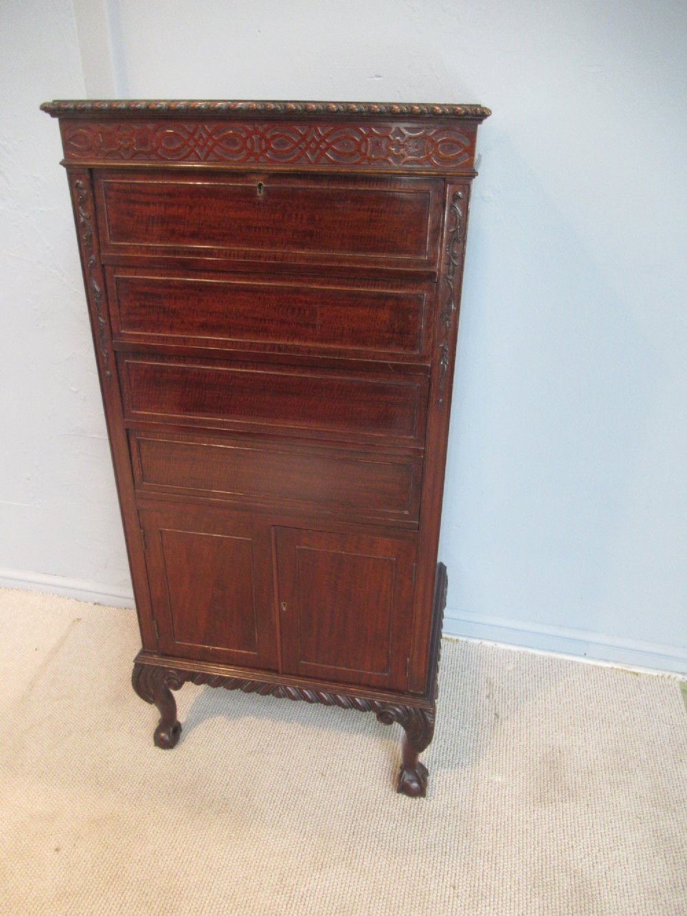 A tall, narrow dark mahogany music cabinet featuring fall-front drawer compartments, carved geometric fretwork, and cabriole legs ending in ball-and-claw feet.