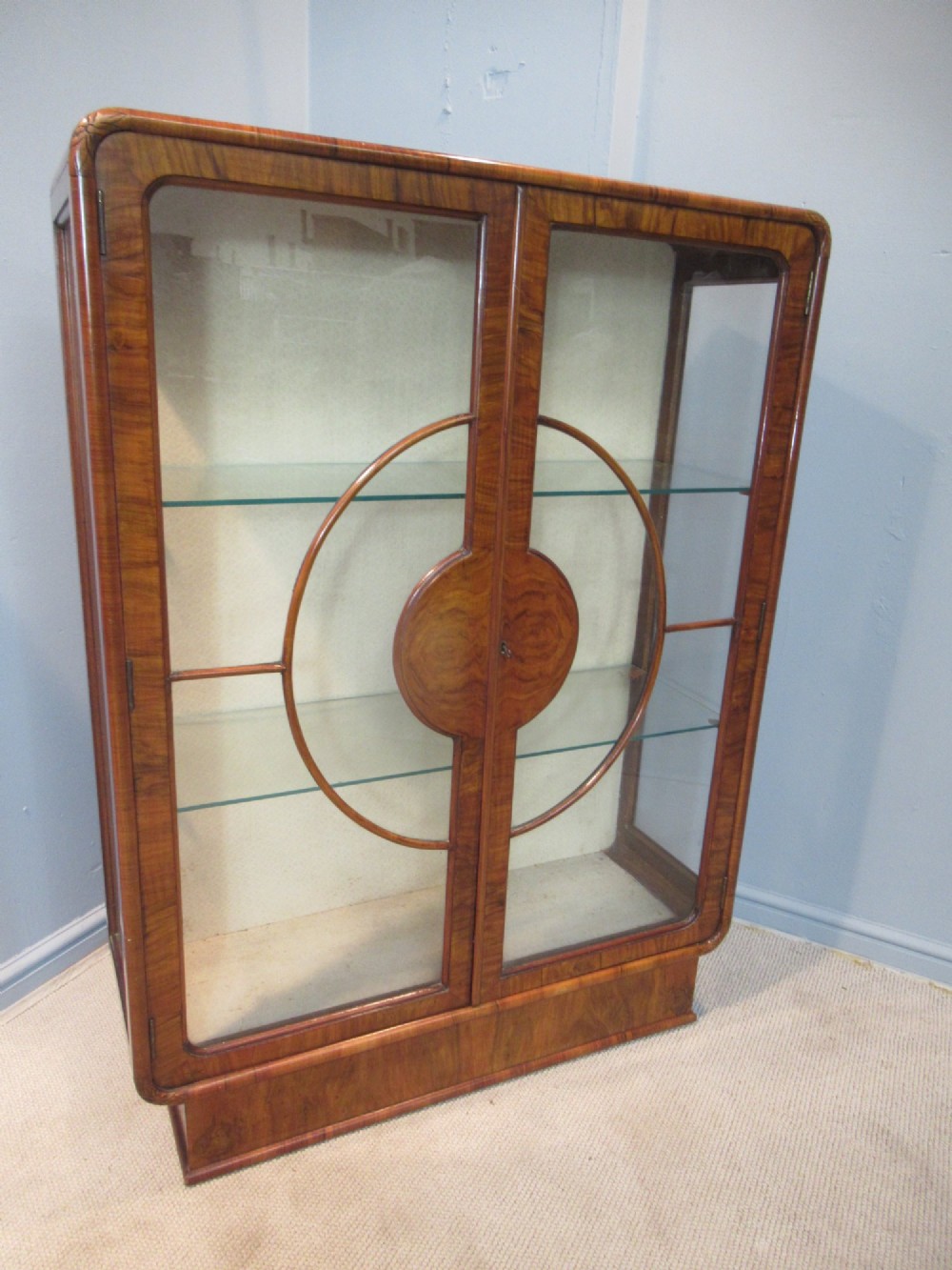 An Art Deco burr walnut display cabinet featuring rounded upper corners, glazed double doors with a central circular wooden motif, and interior glass shelves.