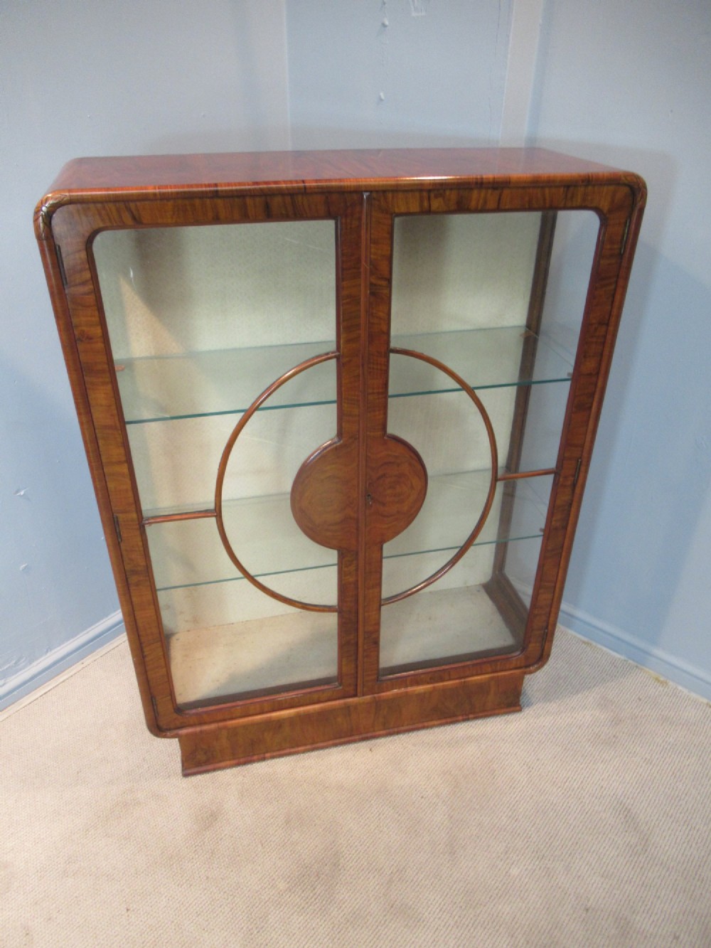 An Art Deco burr walnut display cabinet featuring rounded upper corners, glazed double doors with a central circular wooden motif, and interior glass shelves. View II