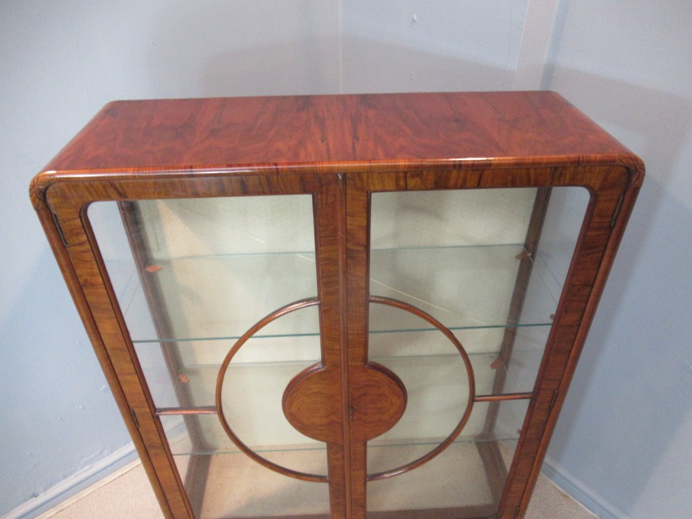 An Art Deco burr walnut display cabinet featuring rounded upper corners, glazed double doors with a central circular wooden motif, and interior glass shelves. View III