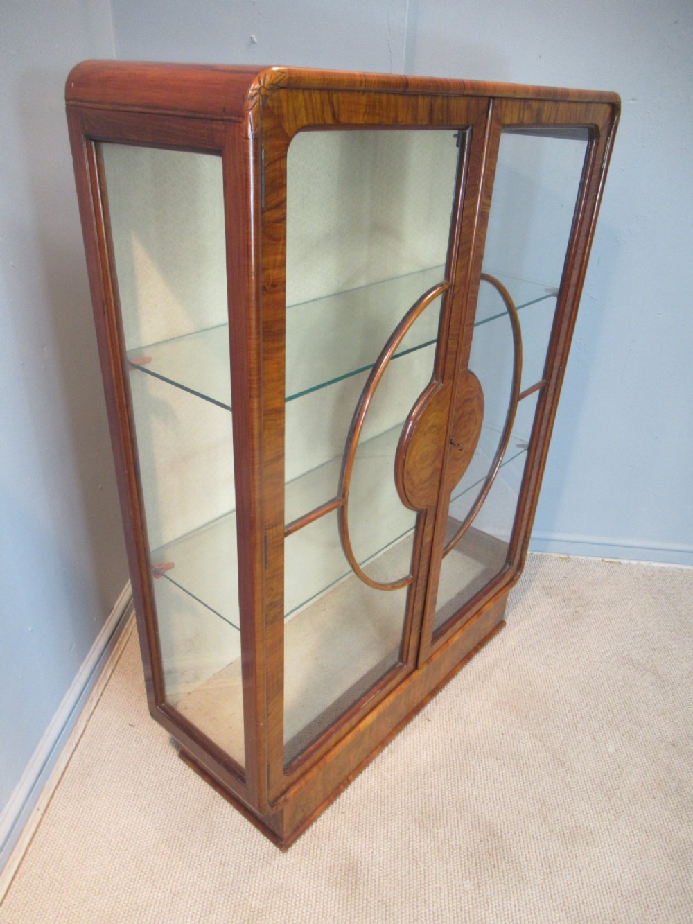 An Art Deco burr walnut display cabinet featuring rounded upper corners, glazed double doors with a central circular wooden motif, and interior glass shelves. View IV