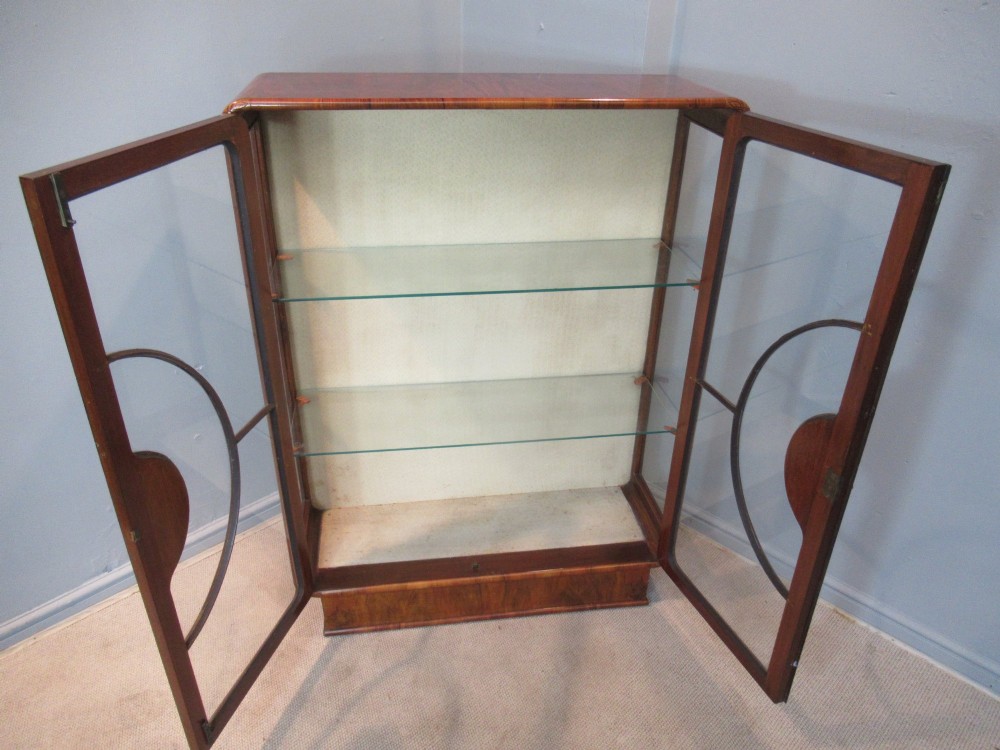 An Art Deco burr walnut display cabinet featuring rounded upper corners, glazed double doors with a central circular wooden motif, and interior glass shelves. View V
