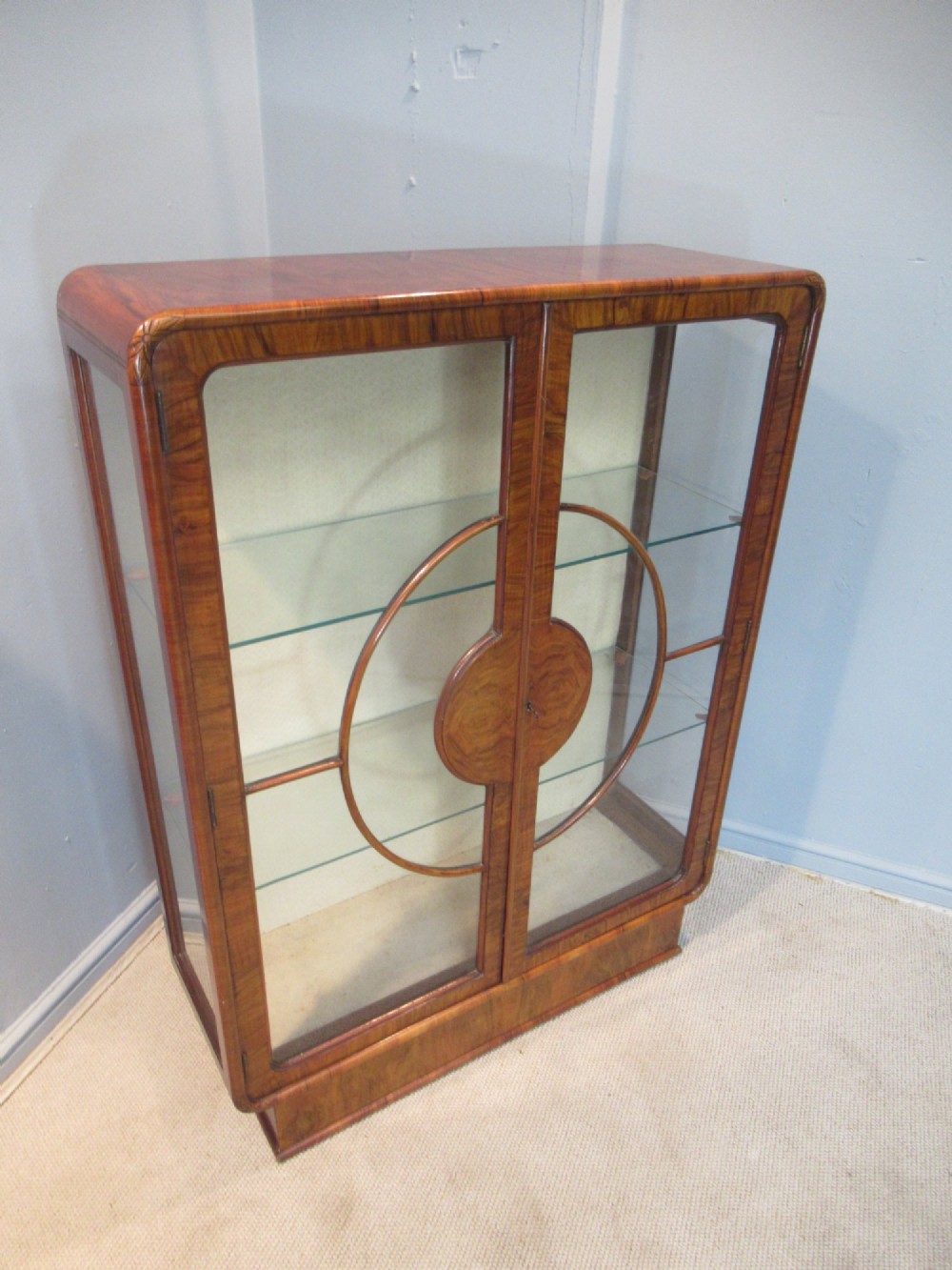 An Art Deco burr walnut display cabinet featuring rounded upper corners, glazed double doors with a central circular wooden motif, and interior glass shelves. View VII