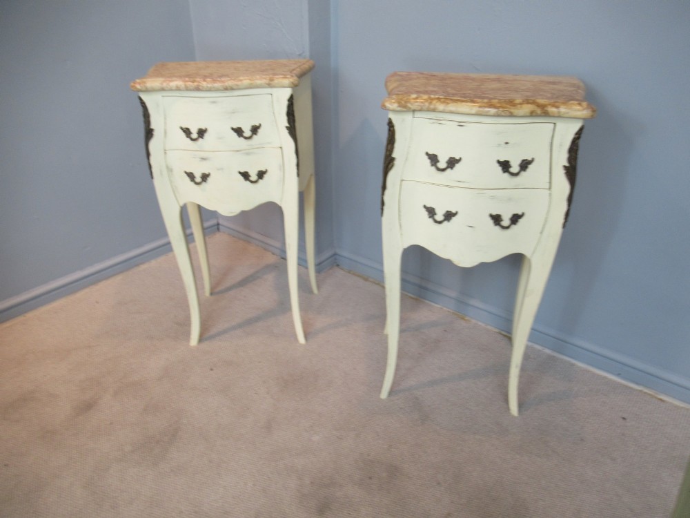A pair of cream painted French style bedside cabinets featuring variegated marble tops, two drawers with brass handles, and slender cabriole legs.