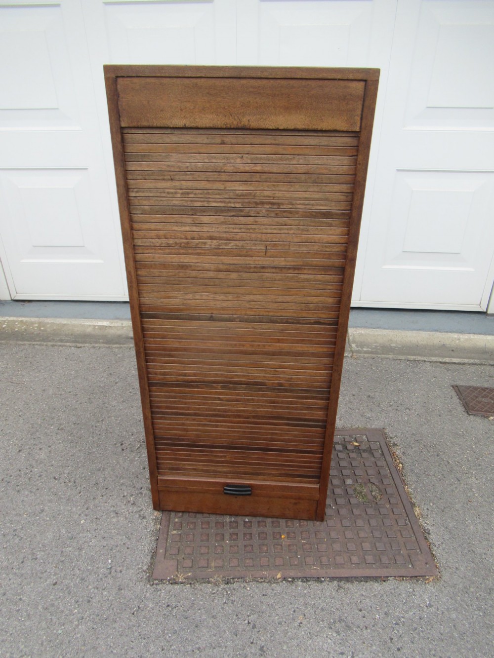 A tall, vertical mahogany filing cabinet with a closed flexible slat tambour front door and a dark handle at the bottom base.