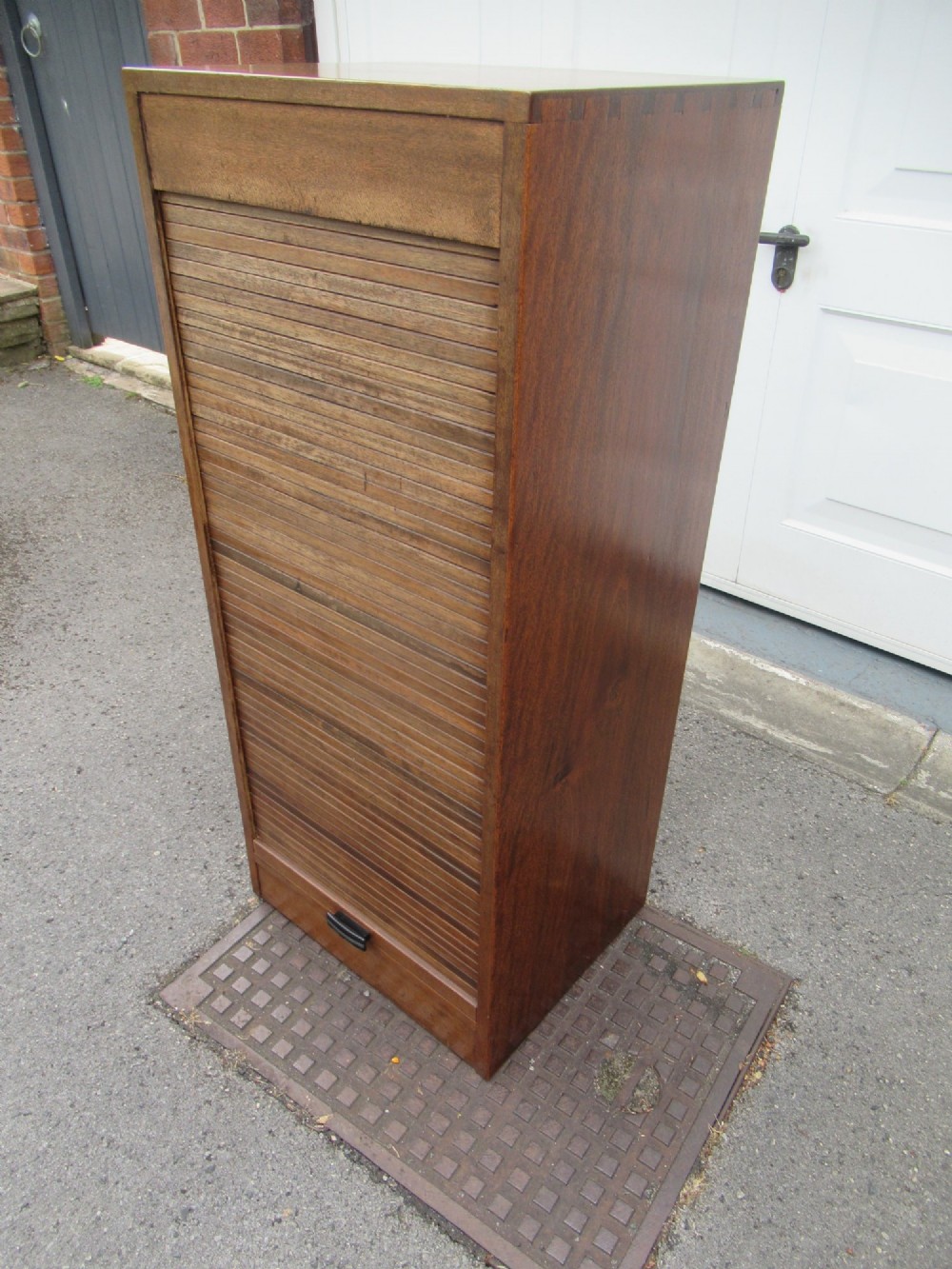 A tall, vertical mahogany filing cabinet with a closed flexible slat tambour front door and a dark handle at the bottom base. View II