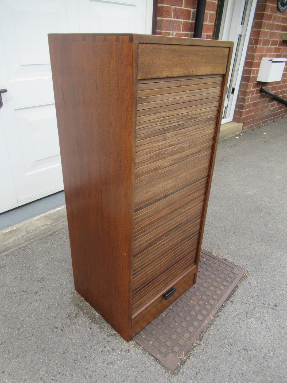 A tall, vertical mahogany filing cabinet with a closed flexible slat tambour front door and a dark handle at the bottom base. View III