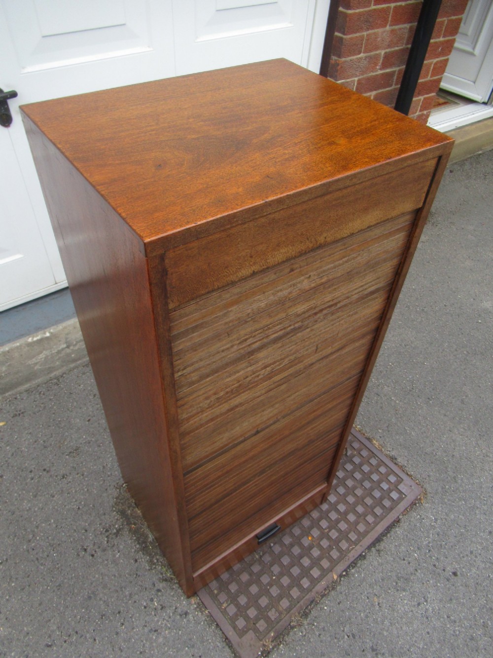 A tall, vertical mahogany filing cabinet with a closed flexible slat tambour front door and a dark handle at the bottom base. View IV