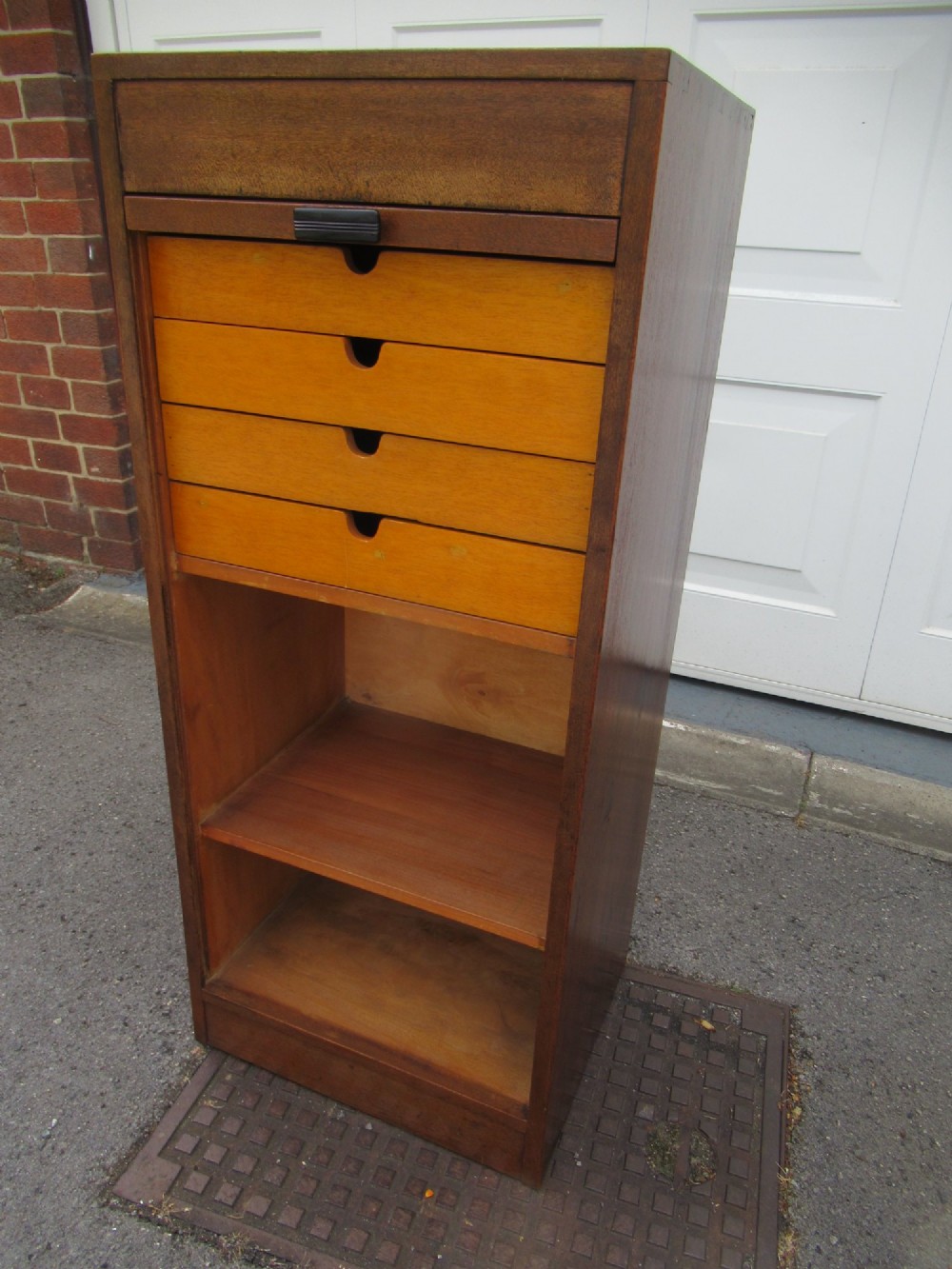 A tall, vertical mahogany filing cabinet with a closed flexible slat tambour front door and a dark handle at the bottom base. View VII