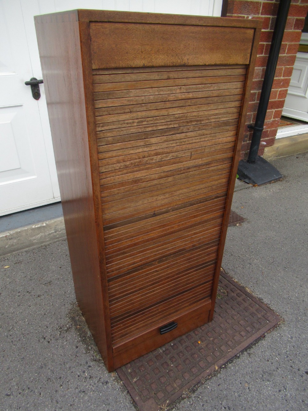 A tall, vertical mahogany filing cabinet with a closed flexible slat tambour front door and a dark handle at the bottom base. View X