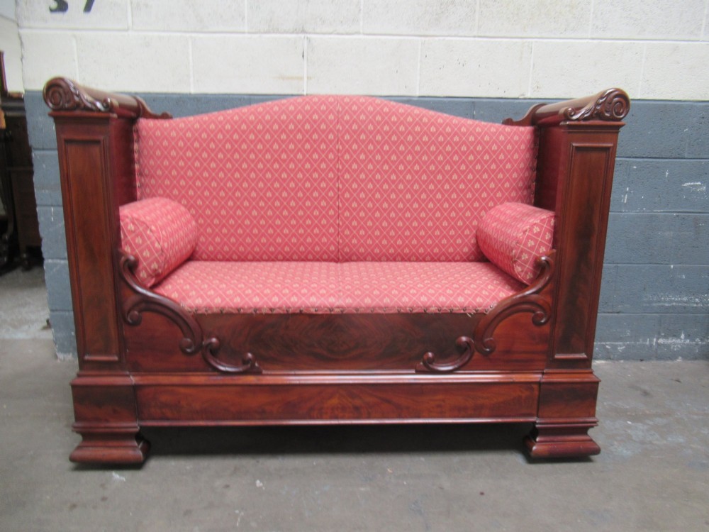 A 19th-century mahogany upholstered bench featuring scrolled armrests, flame mahogany veneered panelling, and red patterned fabric seating with bolster cushions.