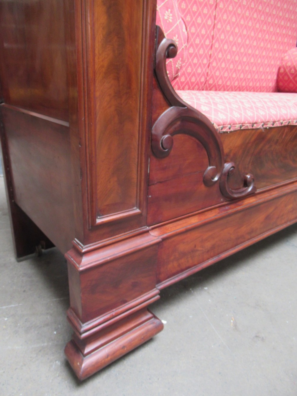 A 19th-century mahogany upholstered bench featuring scrolled armrests, flame mahogany veneered panelling, and red patterned fabric seating with bolster cushions. View V