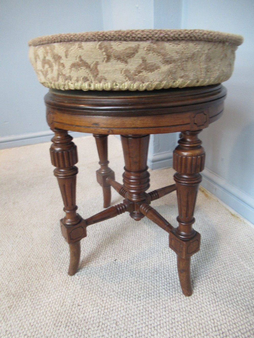 A Victorian walnut revolving piano stool featuring fluted carved legs, cross-stretchers, and a circular padded tapestry upholstered seat. View VI