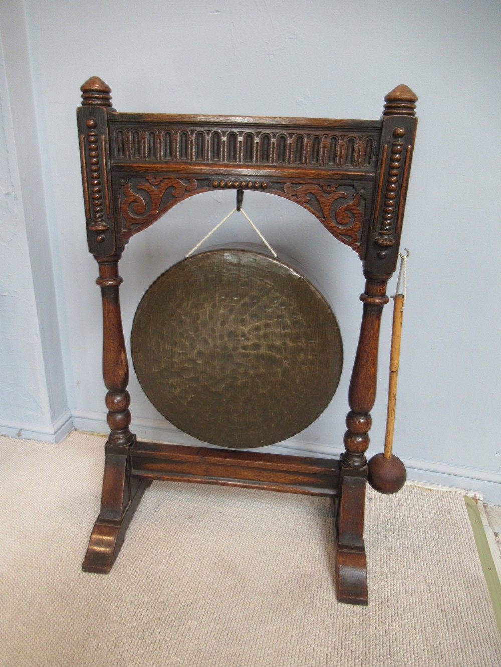 An Edwardian dark oak framed dinner gong featuring a carved arch, turned columns, a suspended circular hammered brass disc, and an attached mallet.