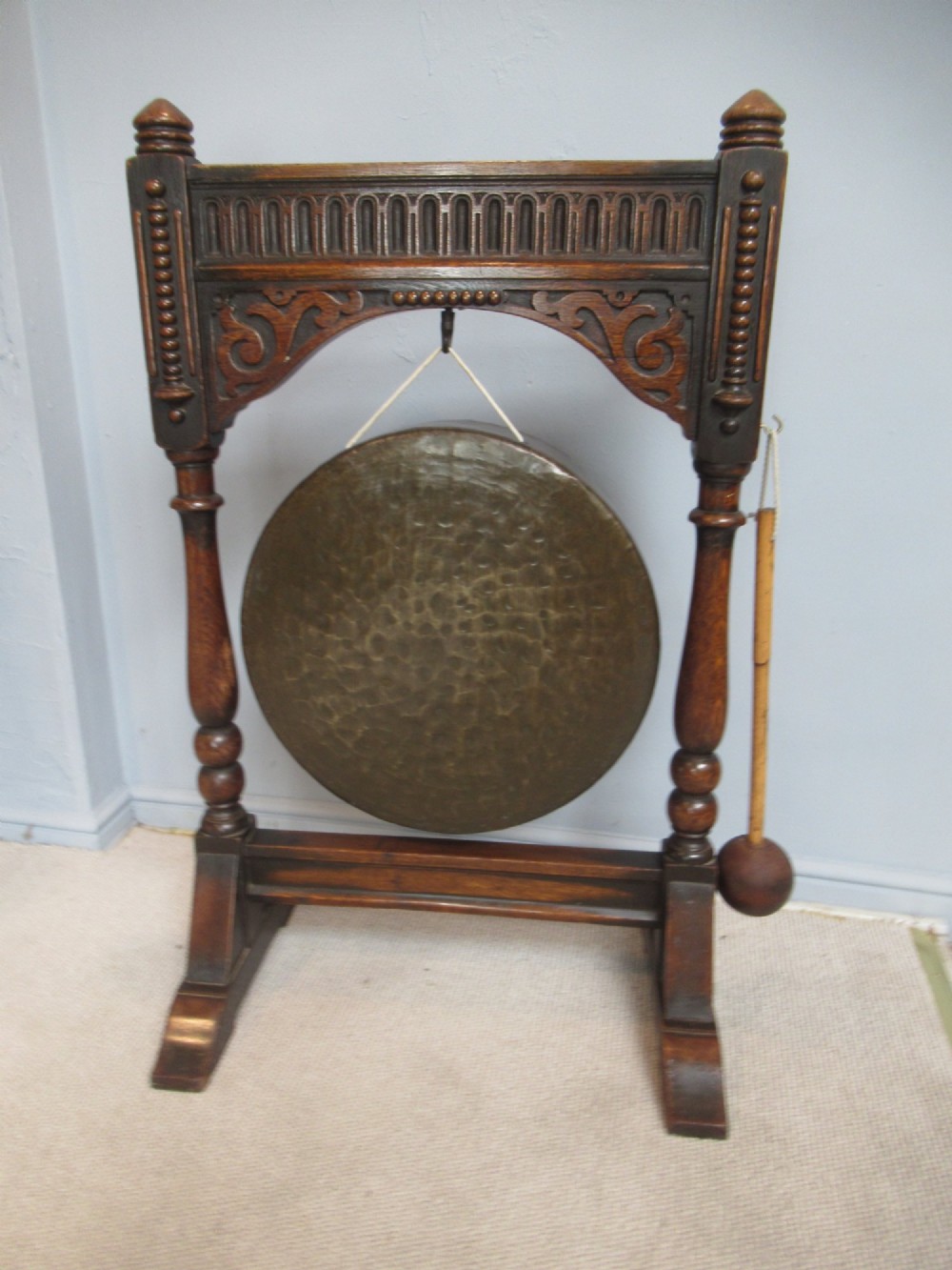 An Edwardian dark oak framed dinner gong featuring a carved arch, turned columns, a suspended circular hammered brass disc, and an attached mallet. View II