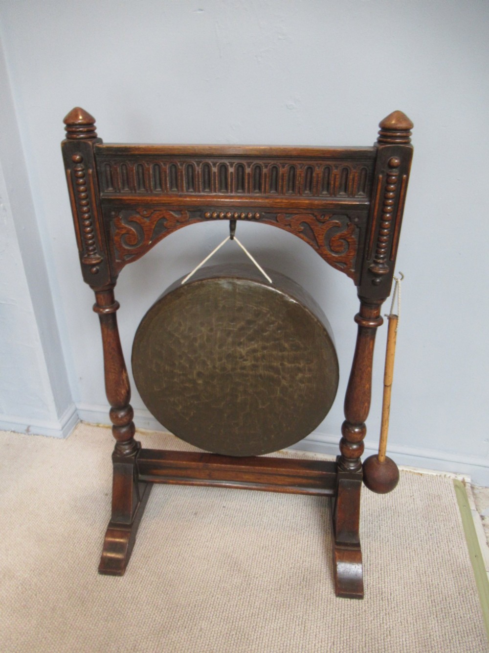 An Edwardian dark oak framed dinner gong featuring a carved arch, turned columns, a suspended circular hammered brass disc, and an attached mallet. View III