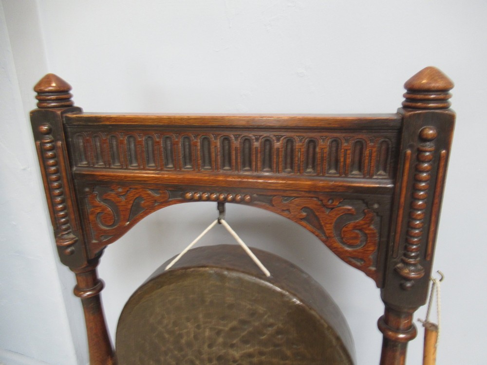 An Edwardian dark oak framed dinner gong featuring a carved arch, turned columns, a suspended circular hammered brass disc, and an attached mallet. View IV