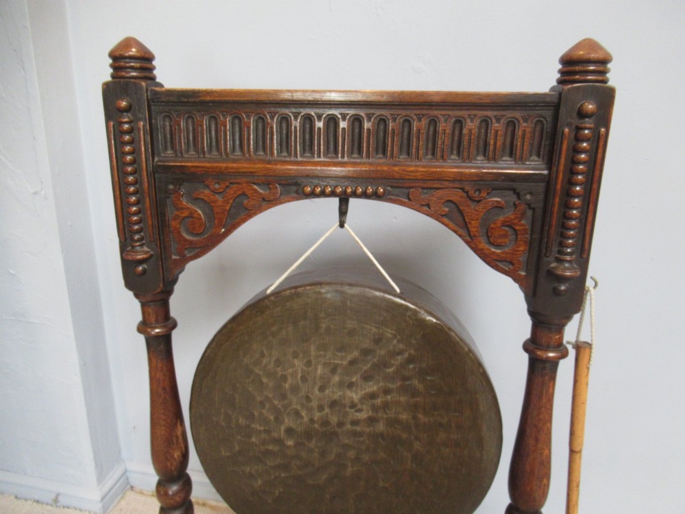 An Edwardian dark oak framed dinner gong featuring a carved arch, turned columns, a suspended circular hammered brass disc, and an attached mallet. View V