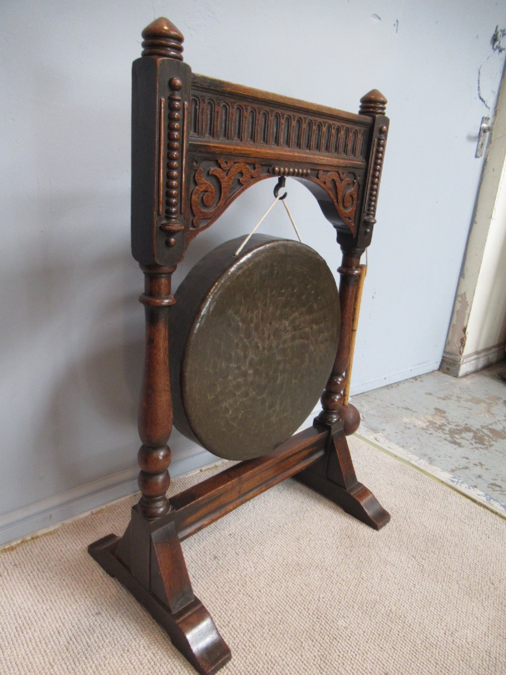An Edwardian dark oak framed dinner gong featuring a carved arch, turned columns, a suspended circular hammered brass disc, and an attached mallet. View VII
