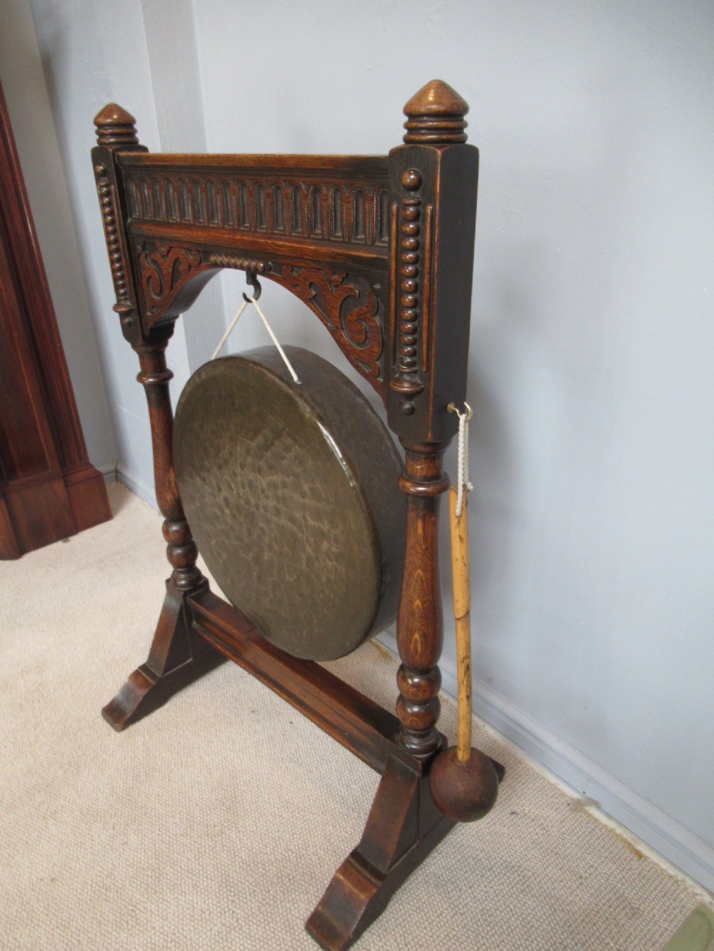 An Edwardian dark oak framed dinner gong featuring a carved arch, turned columns, a suspended circular hammered brass disc, and an attached mallet. View VIII
