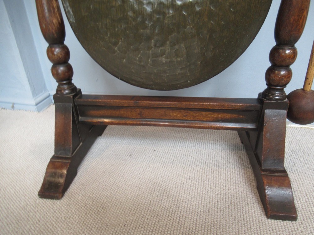 An Edwardian dark oak framed dinner gong featuring a carved arch, turned columns, a suspended circular hammered brass disc, and an attached mallet. View IX