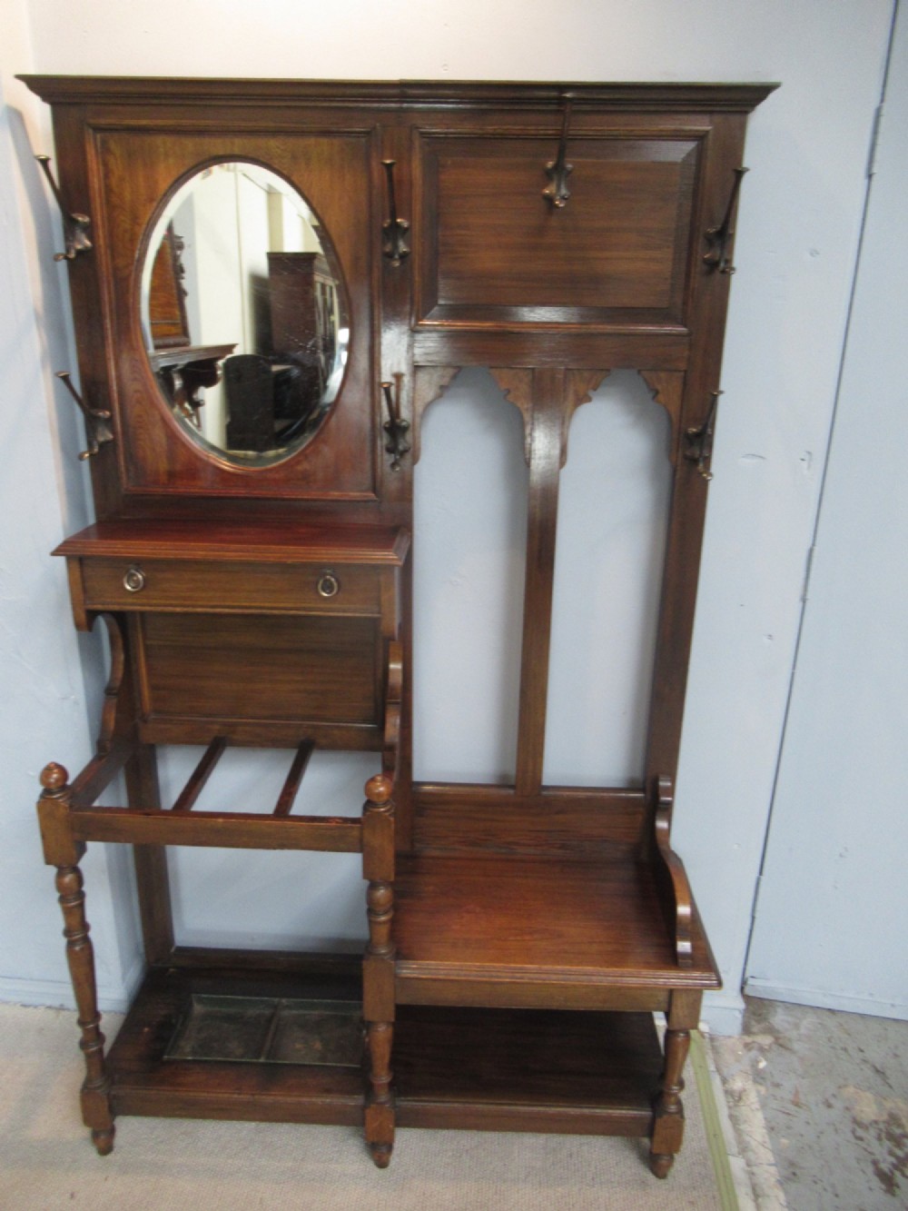 An Edwardian dark oak hall stand featuring an oval mirror, small glove drawer, integrated seat, stick holder, and multiple brass coat hooks. View II