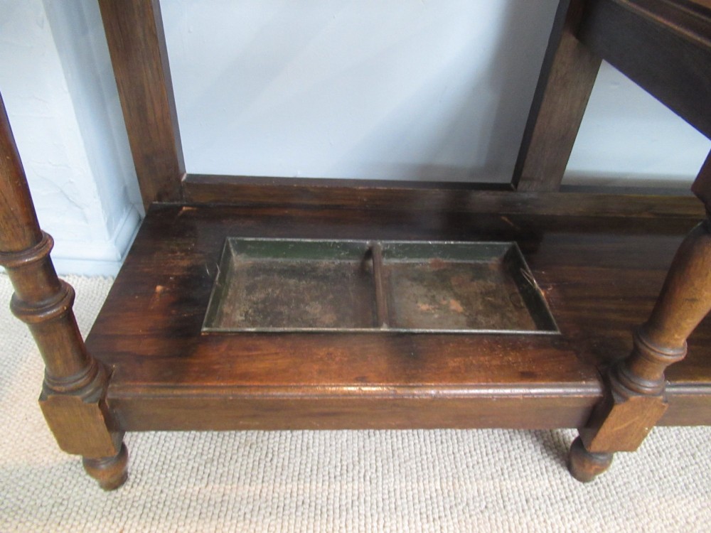 An Edwardian dark oak hall stand featuring an oval mirror, small glove drawer, integrated seat, stick holder, and multiple brass coat hooks. View IV