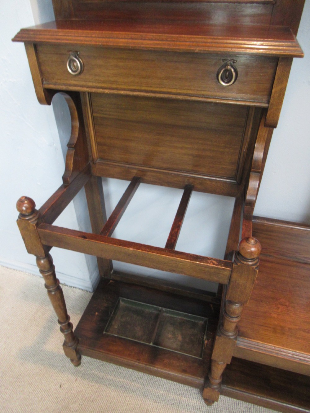 An Edwardian dark oak hall stand featuring an oval mirror, small glove drawer, integrated seat, stick holder, and multiple brass coat hooks. View V