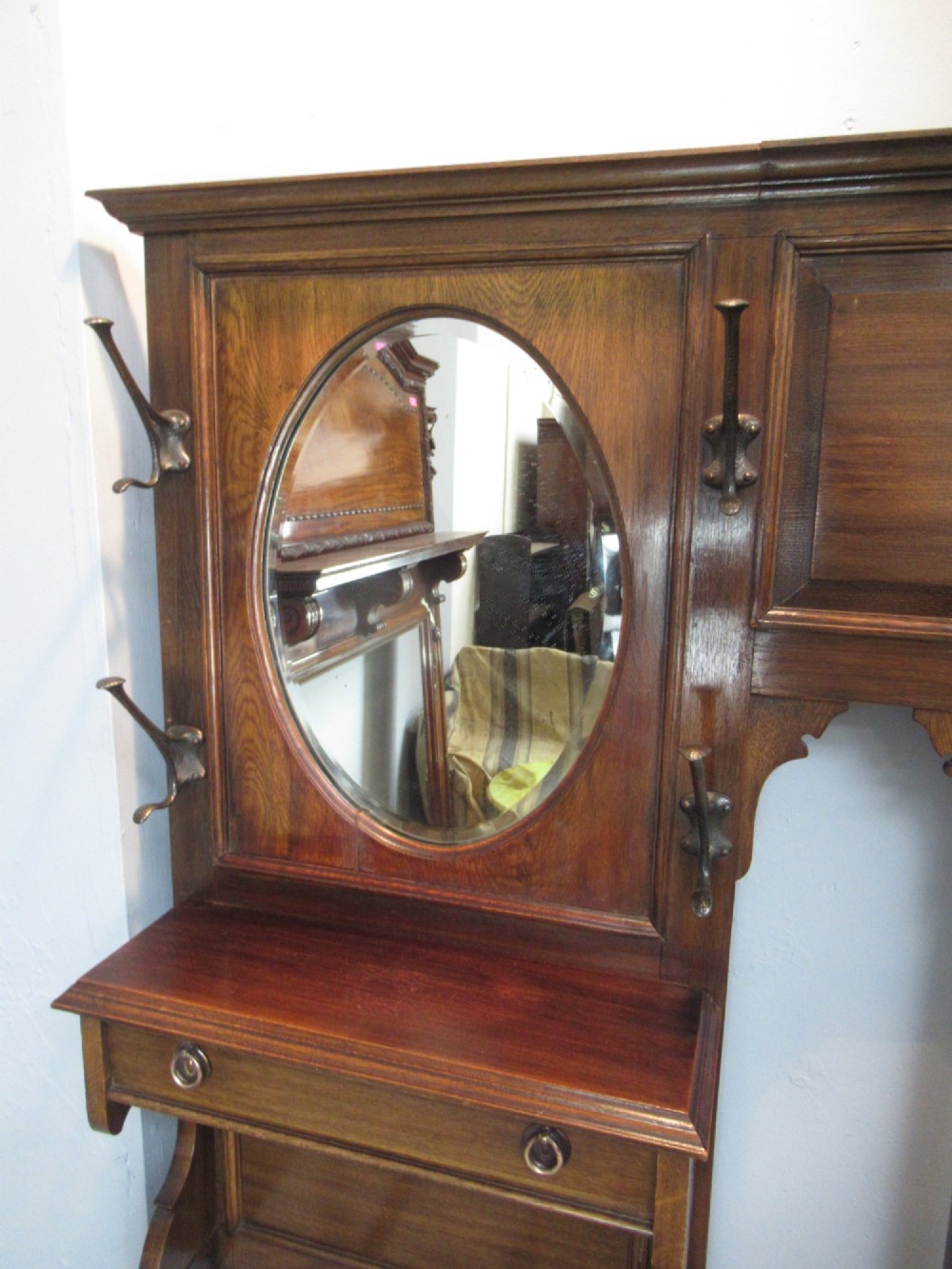 An Edwardian dark oak hall stand featuring an oval mirror, small glove drawer, integrated seat, stick holder, and multiple brass coat hooks. View VI