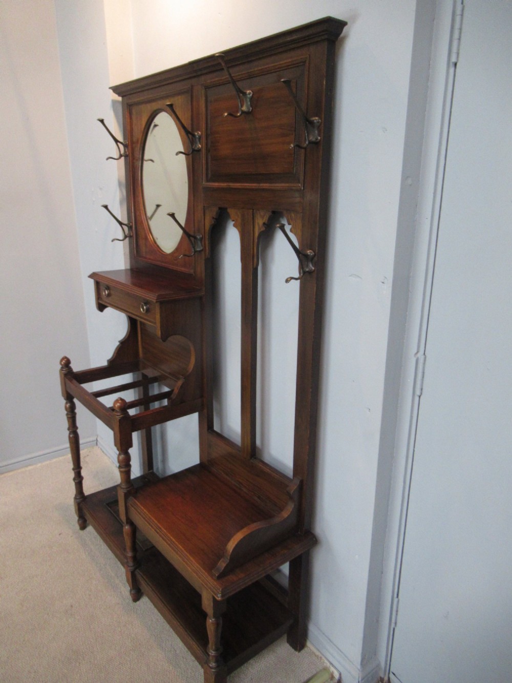 An Edwardian dark oak hall stand featuring an oval mirror, small glove drawer, integrated seat, stick holder, and multiple brass coat hooks. View VII