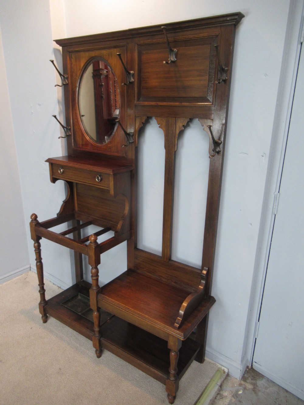 An Edwardian dark oak hall stand featuring an oval mirror, small glove drawer, integrated seat, stick holder, and multiple brass coat hooks. View VIII