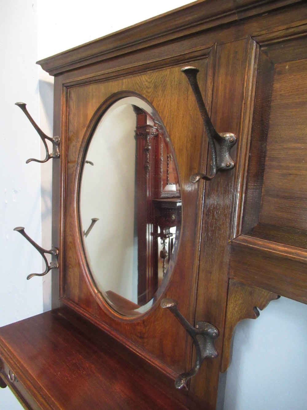 An Edwardian dark oak hall stand featuring an oval mirror, small glove drawer, integrated seat, stick holder, and multiple brass coat hooks. View IX