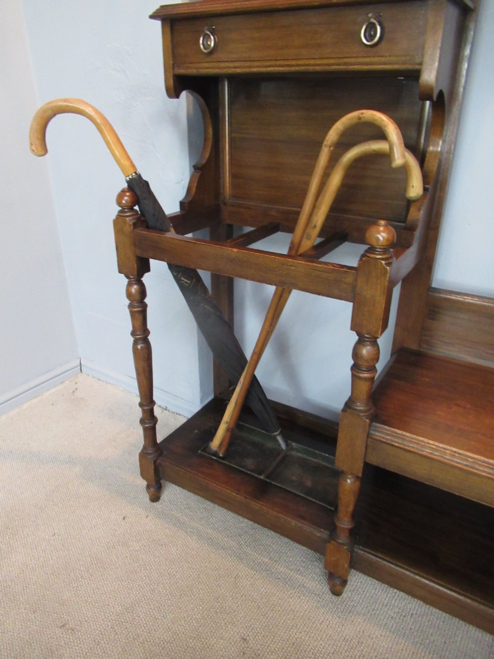 An Edwardian dark oak hall stand featuring an oval mirror, small glove drawer, integrated seat, stick holder, and multiple brass coat hooks. View X
