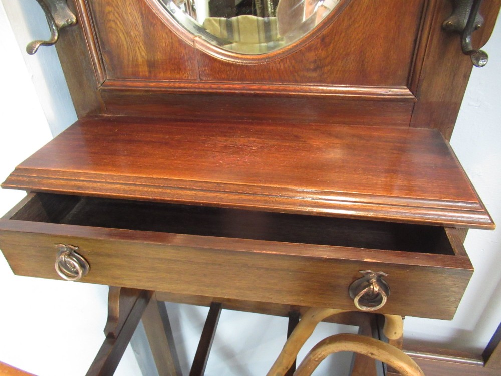 An Edwardian dark oak hall stand featuring an oval mirror, small glove drawer, integrated seat, stick holder, and multiple brass coat hooks. View XI