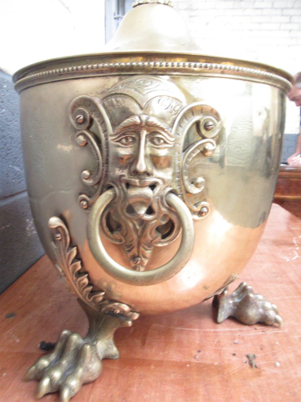 A polished brass lidded decorative bucket with a finial handle, lion mask side rings, and ornate claw feet. View II