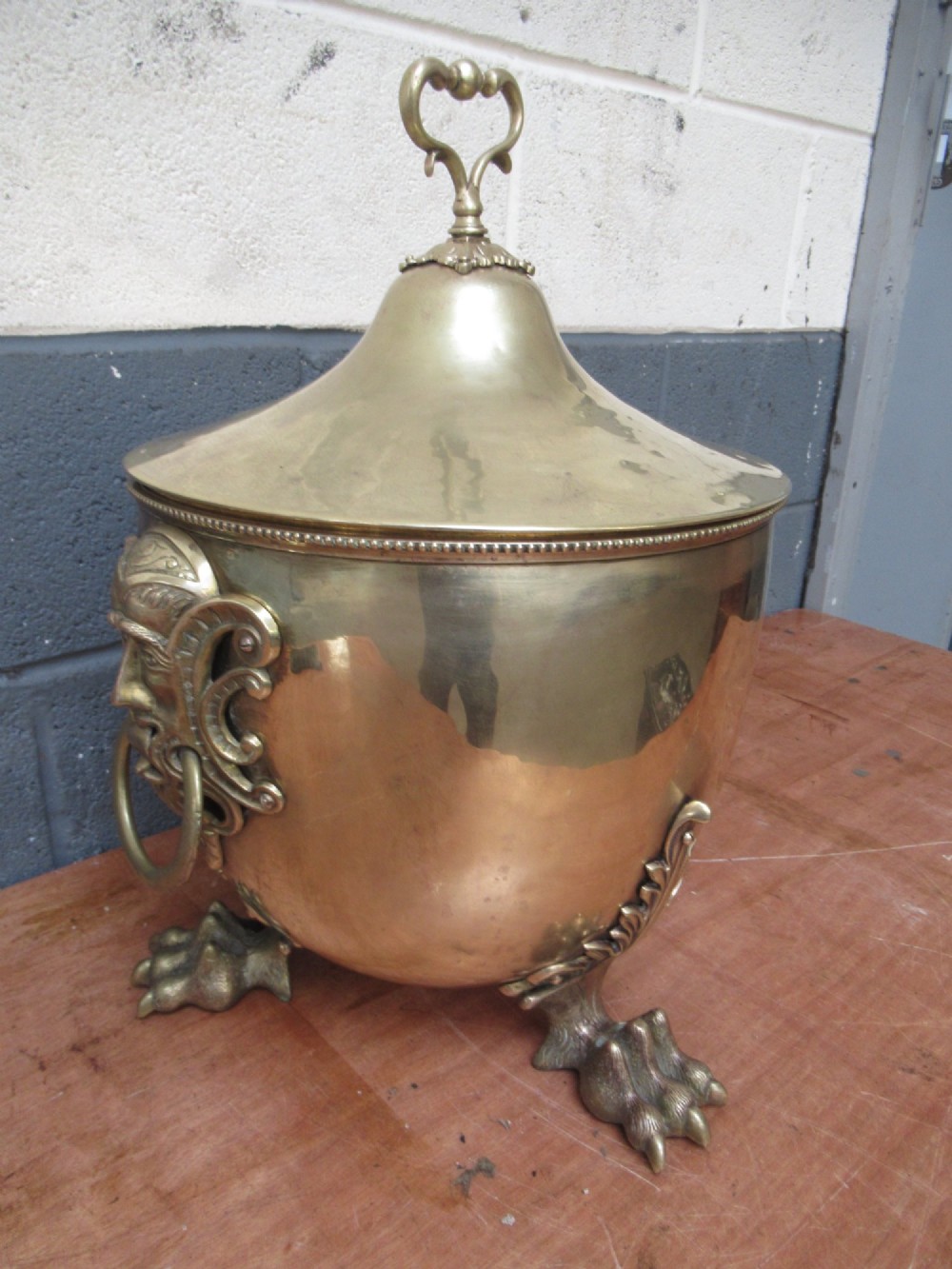 A polished brass lidded decorative bucket with a finial handle, lion mask side rings, and ornate claw feet. View V