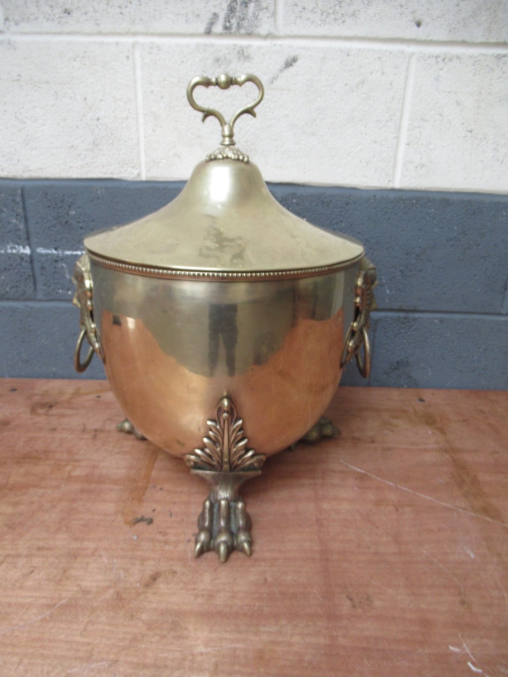A polished brass lidded decorative bucket with a finial handle, lion mask side rings, and ornate claw feet. View X