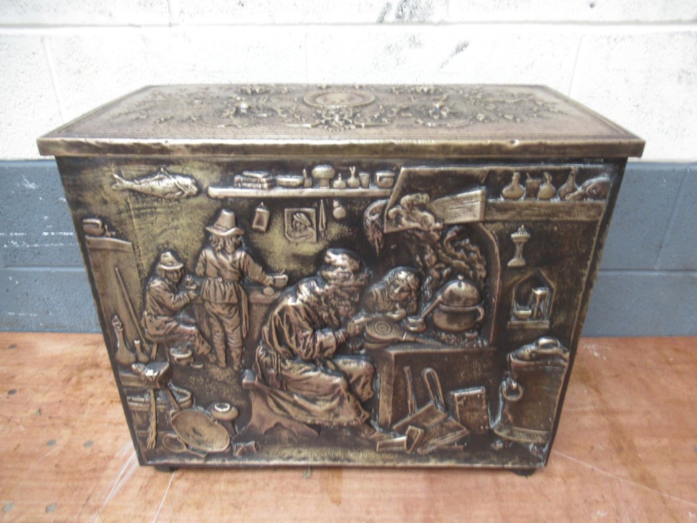 A rectangular Victorian brass log box featuring a detailed embossed relief scene of figures inside a room on the front panel. View II