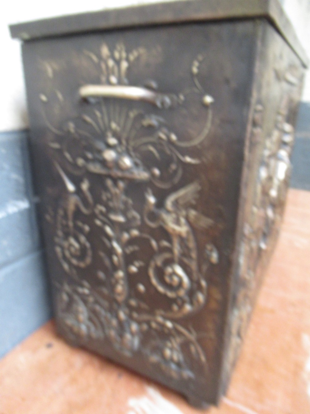 A rectangular Victorian brass log box featuring a detailed embossed relief scene of figures inside a room on the front panel. View IV