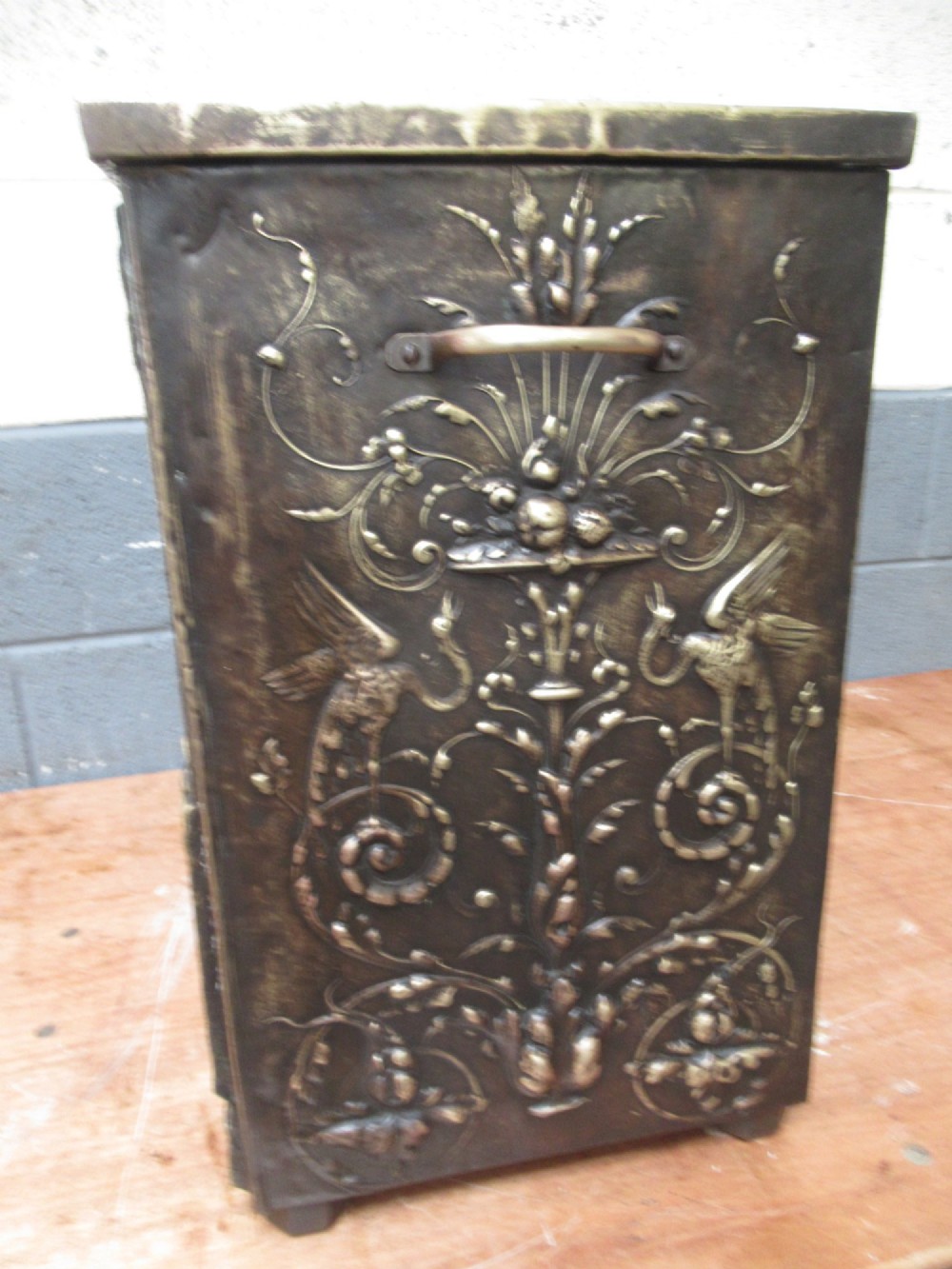 A rectangular Victorian brass log box featuring a detailed embossed relief scene of figures inside a room on the front panel. View V