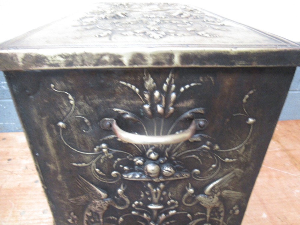 A rectangular Victorian brass log box featuring a detailed embossed relief scene of figures inside a room on the front panel. View VI