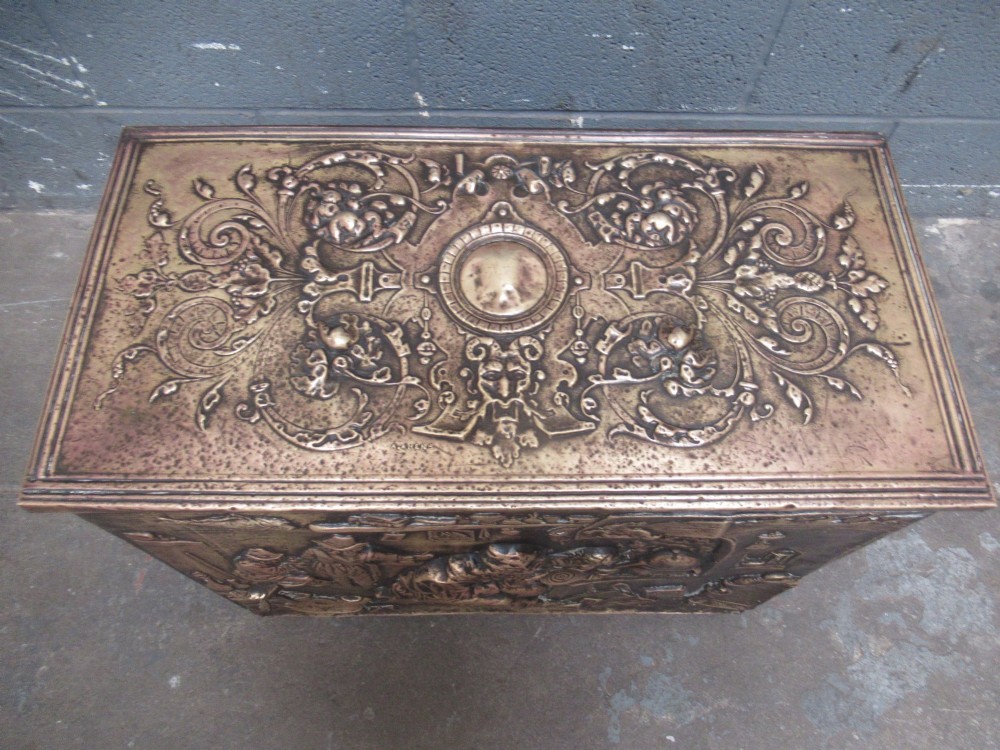 A rectangular Victorian brass log box featuring a detailed embossed relief scene of figures inside a room on the front panel. View VIII