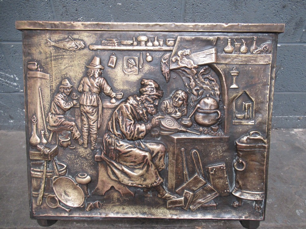 A rectangular Victorian brass log box featuring a detailed embossed relief scene of figures inside a room on the front panel. View X