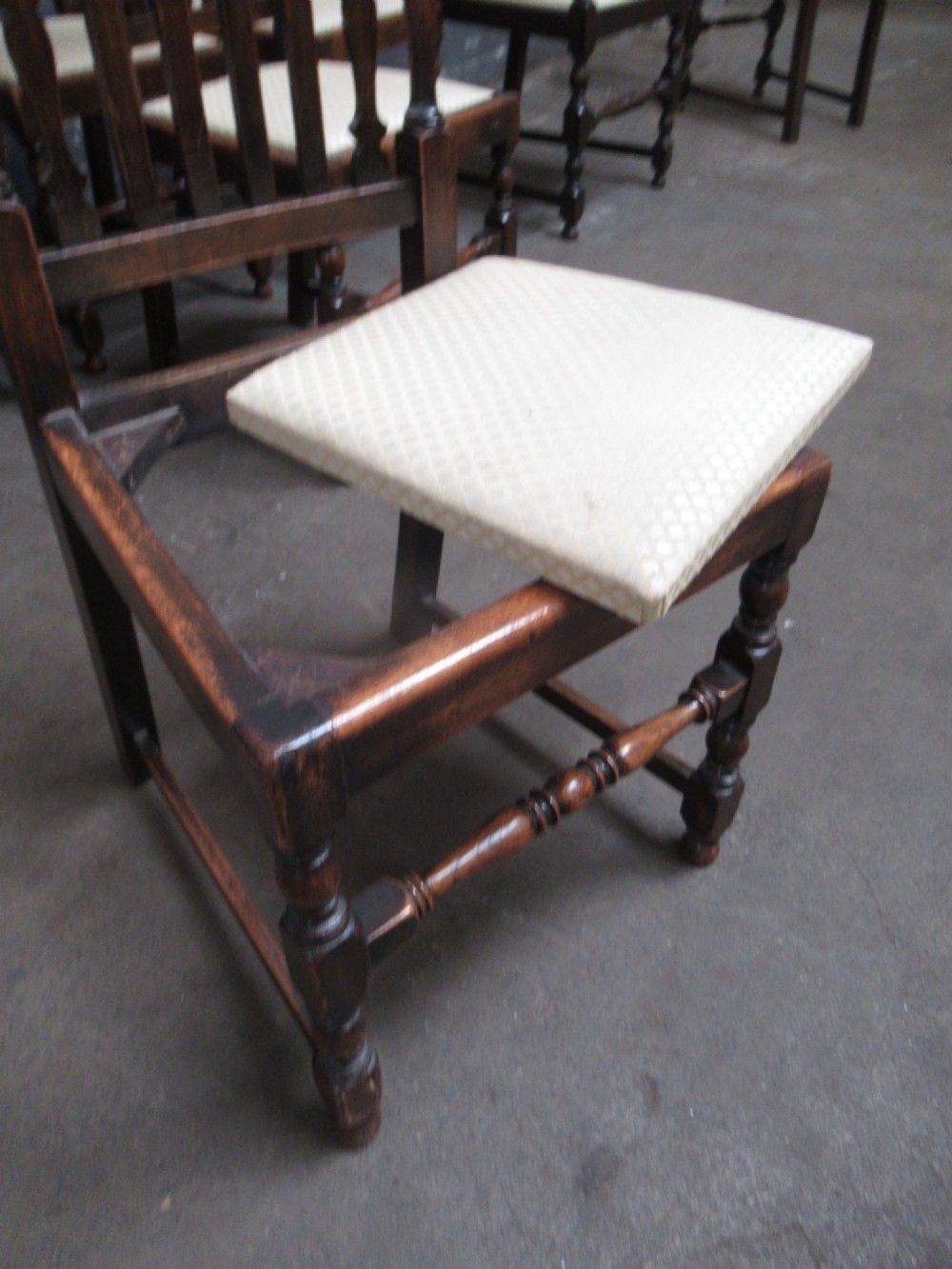 A set of eight 1920s dark oak dining chairs with carved cresting rails, vertical back slats, turned legs, and light cream upholstered seats, including two carvers with armrests. View VII